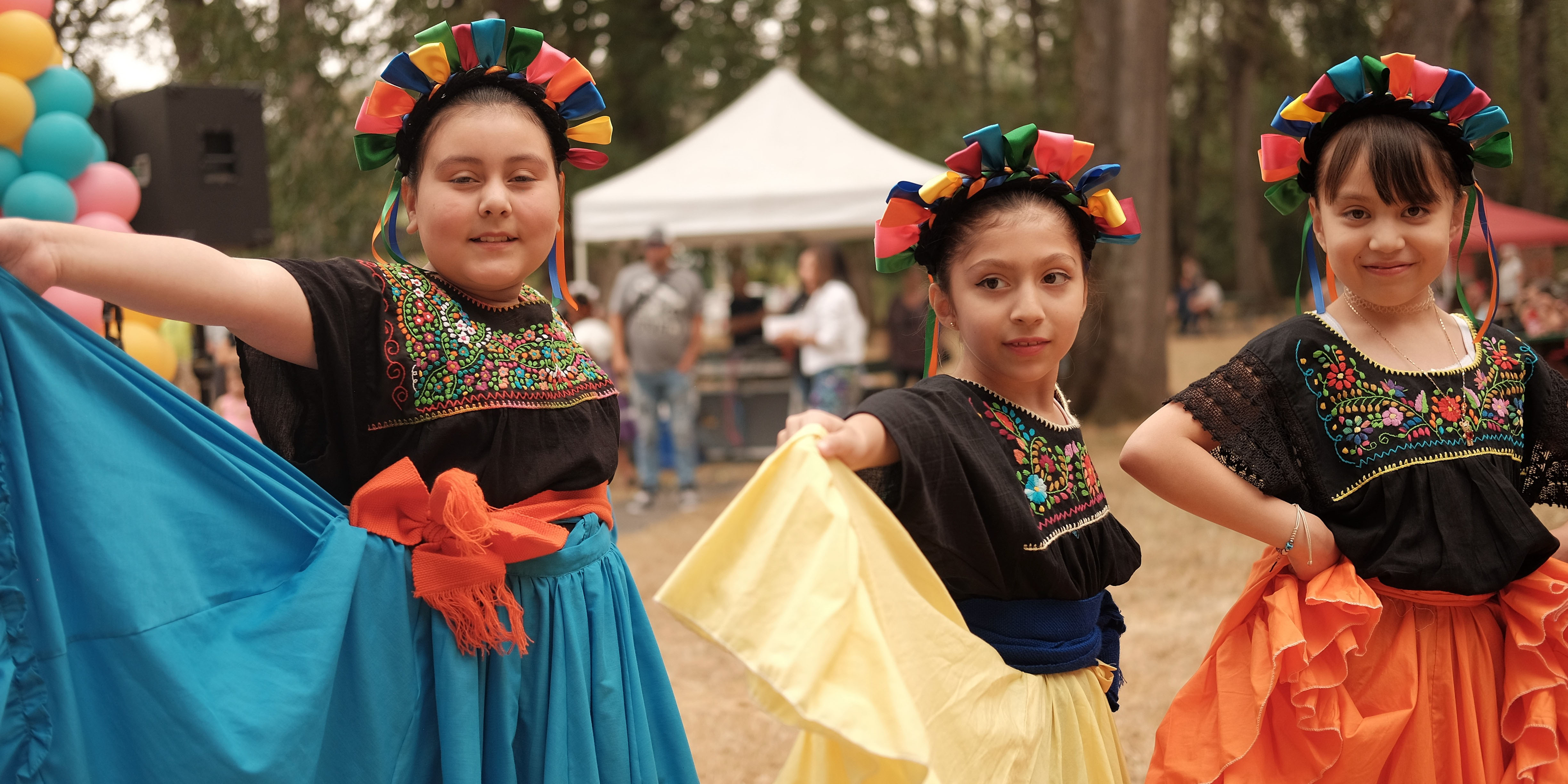 Three girls in folk dresses hold out their bright-colored skirts