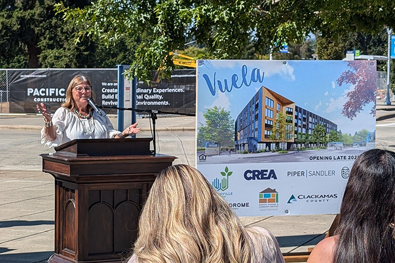 County Commissioner Martha Schrader speaks at the groundbreaking of Vuela in Wilsonville as a bus passes behind her at the Wilsonville Transit Station